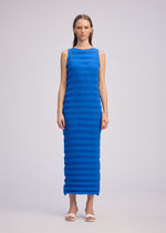 A woman faces forward in the ISSEY MIYAKE SPRING PLEATS sleeveless, bright blue, ankle-length dress with a ribbed texture, paired with white sandals against a plain light gray background.