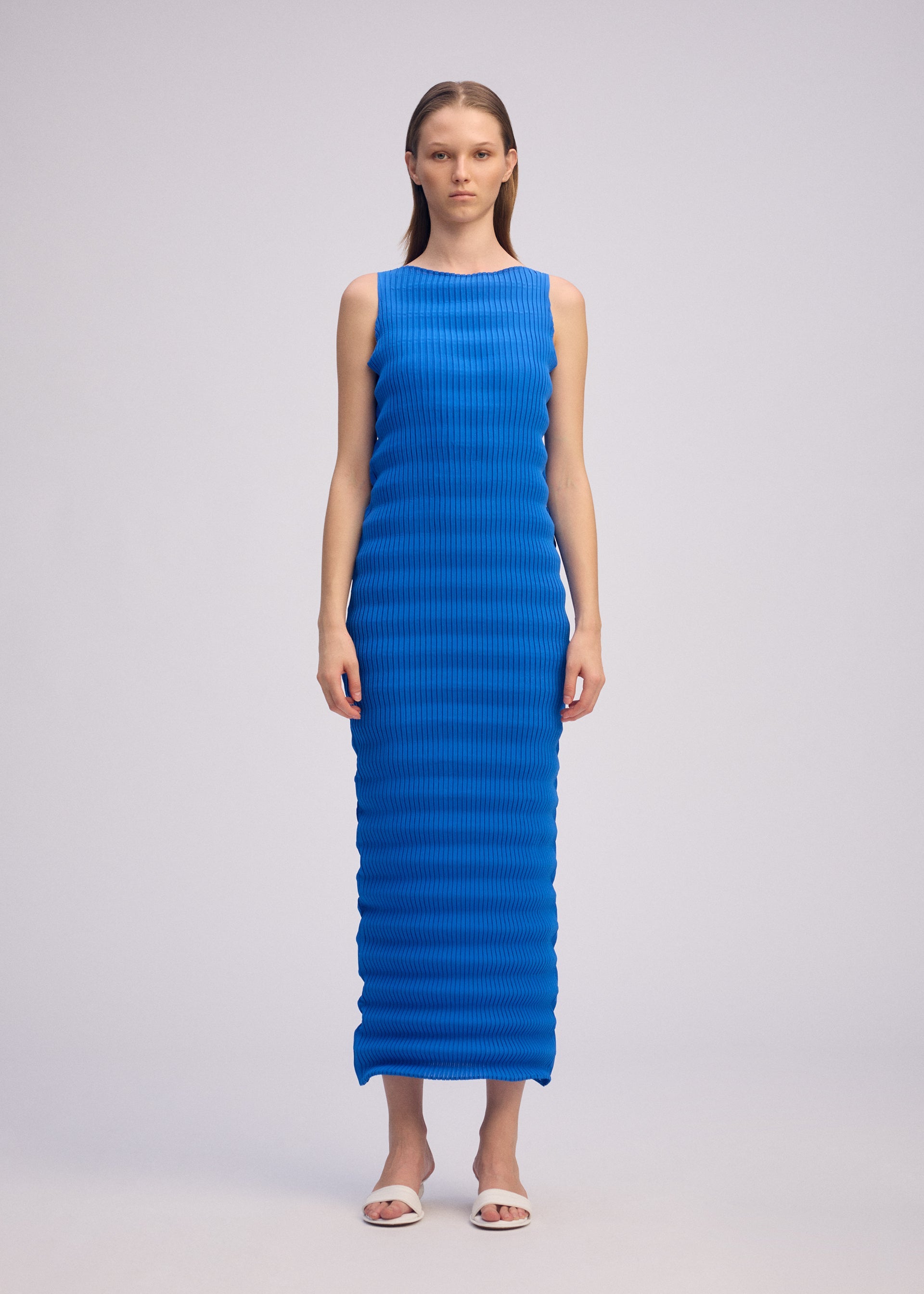 Zoomed view A woman faces forward in the ISSEY MIYAKE SPRING PLEATS sleeveless, bright blue, ankle-length dress with a ribbed texture, paired with white sandals against a plain light gray background.