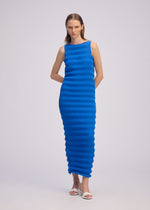 A woman poses against a plain background in the ISSEY MIYAKE SPRING PLEATS sleeveless high-twist polyester dress, featuring a fitted ribbed silhouette in bright blue, paired with white sandals. Her arms rest relaxed behind her back.