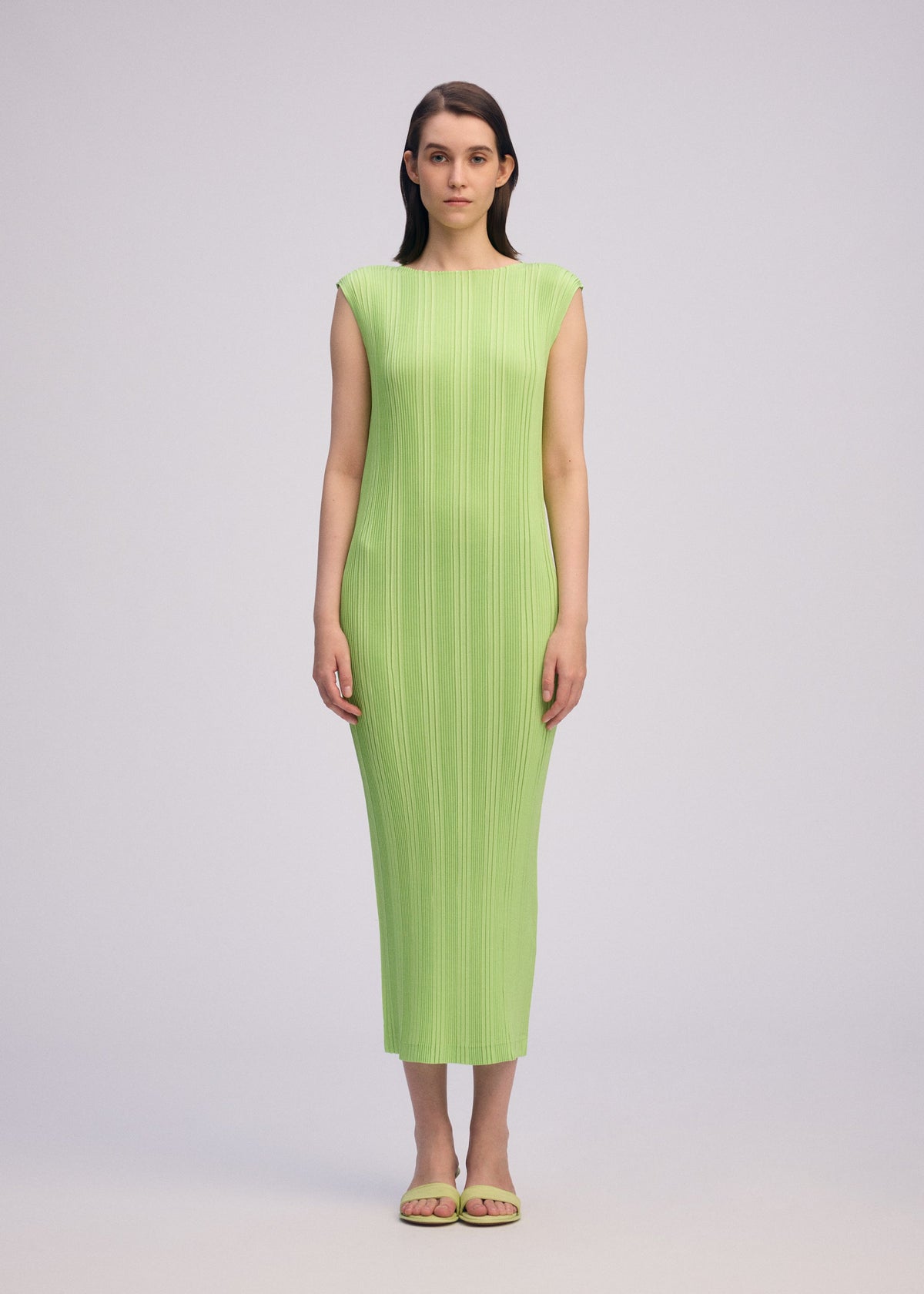 A woman stands against a plain light background, wearing ISSEY MIYAKE’s LINEN LIKE RIB PLEATS sleeveless, form-fitting light green dress with a round neck and pairs it with matching green sandals, facing forward with arms relaxed at her sides. view 5 of 6