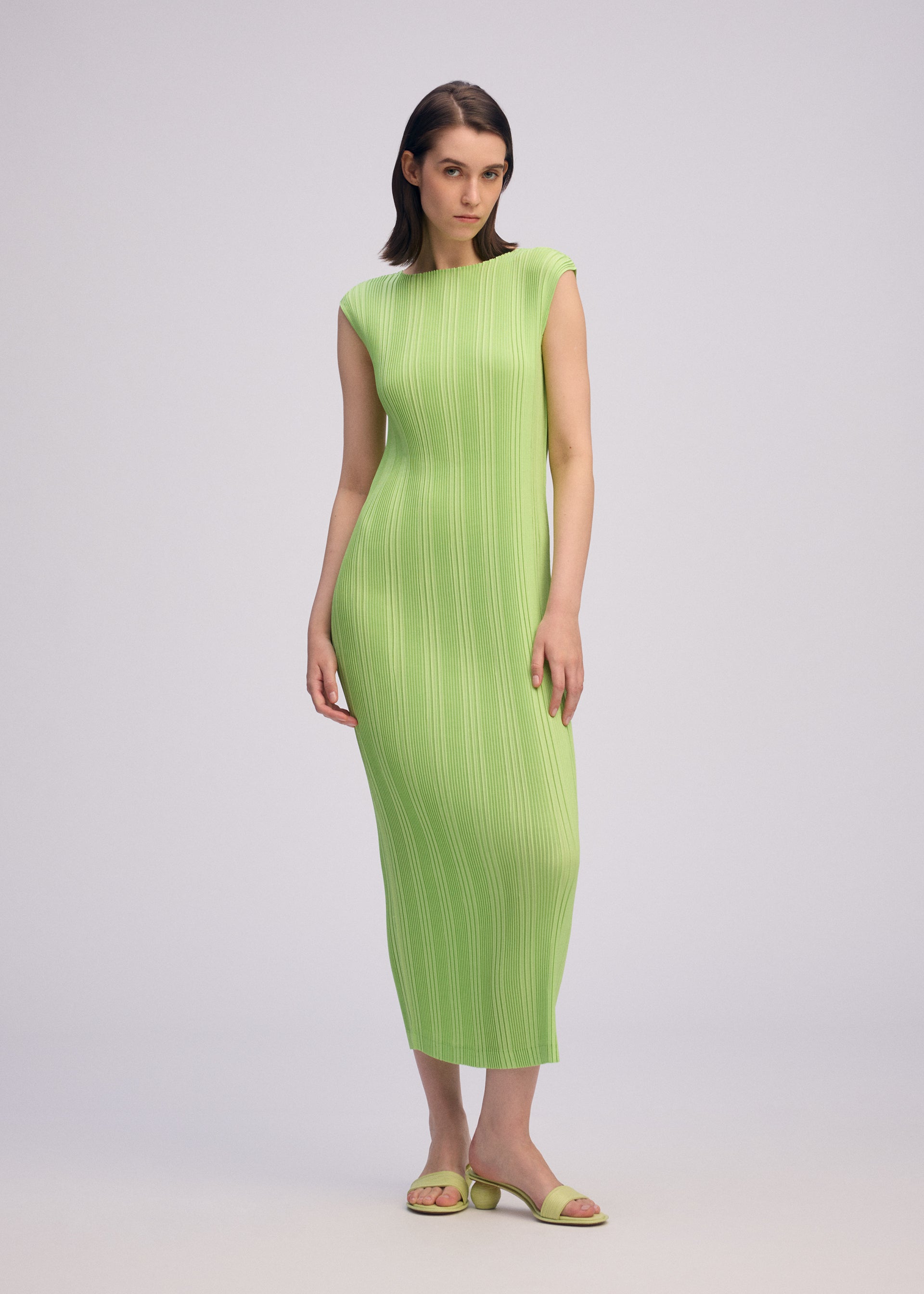 Zoomed view A woman wears ISSEY MIYAKE's LINEN LIKE RIB PLEATS—a fitted, sleeveless light green ankle-length dress—paired with matching sandals. She has straight dark hair and a neutral expression against a plain light background.