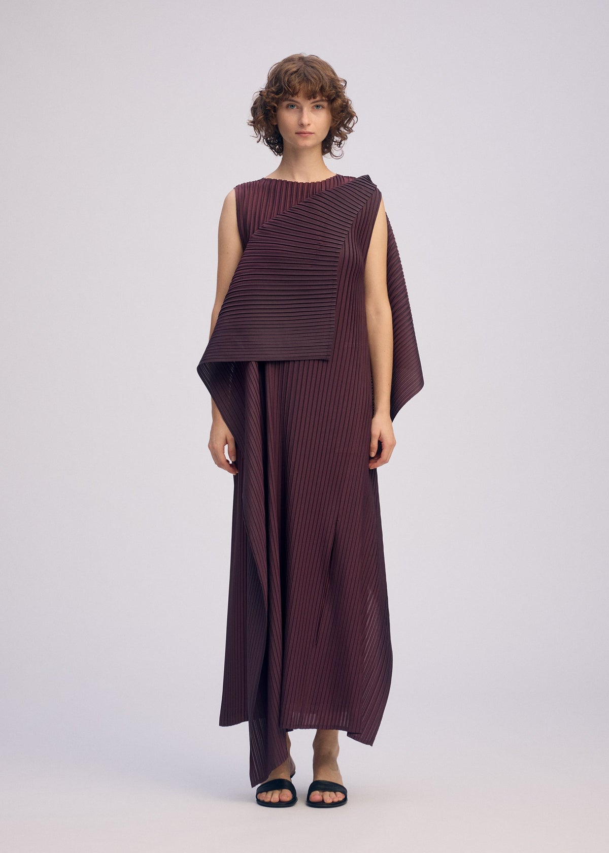 A person with short, curly hair wears the ISSEY MIYAKE PLEATED ON PLEATS sleeveless maroon asymmetrical dress and black sandals, standing against a plain light background. view 5 of 8
