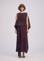 A person with short, curly hair wears the ISSEY MIYAKE PLEATED ON PLEATS sleeveless maroon asymmetrical dress and black sandals, standing against a plain light background.