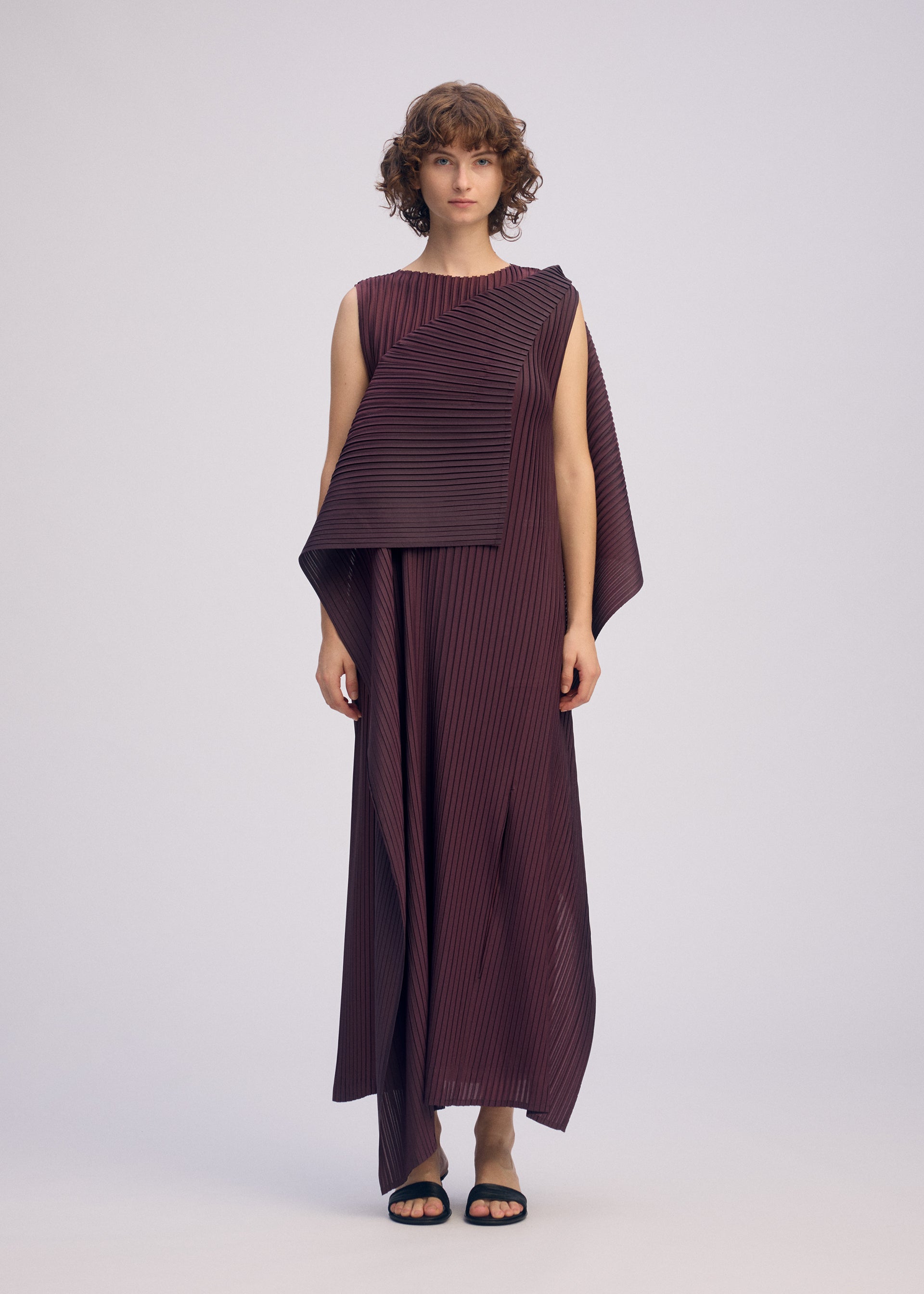 Zoomed view A person with short, curly hair wears the ISSEY MIYAKE PLEATED ON PLEATS sleeveless maroon asymmetrical dress and black sandals, standing against a plain light background.