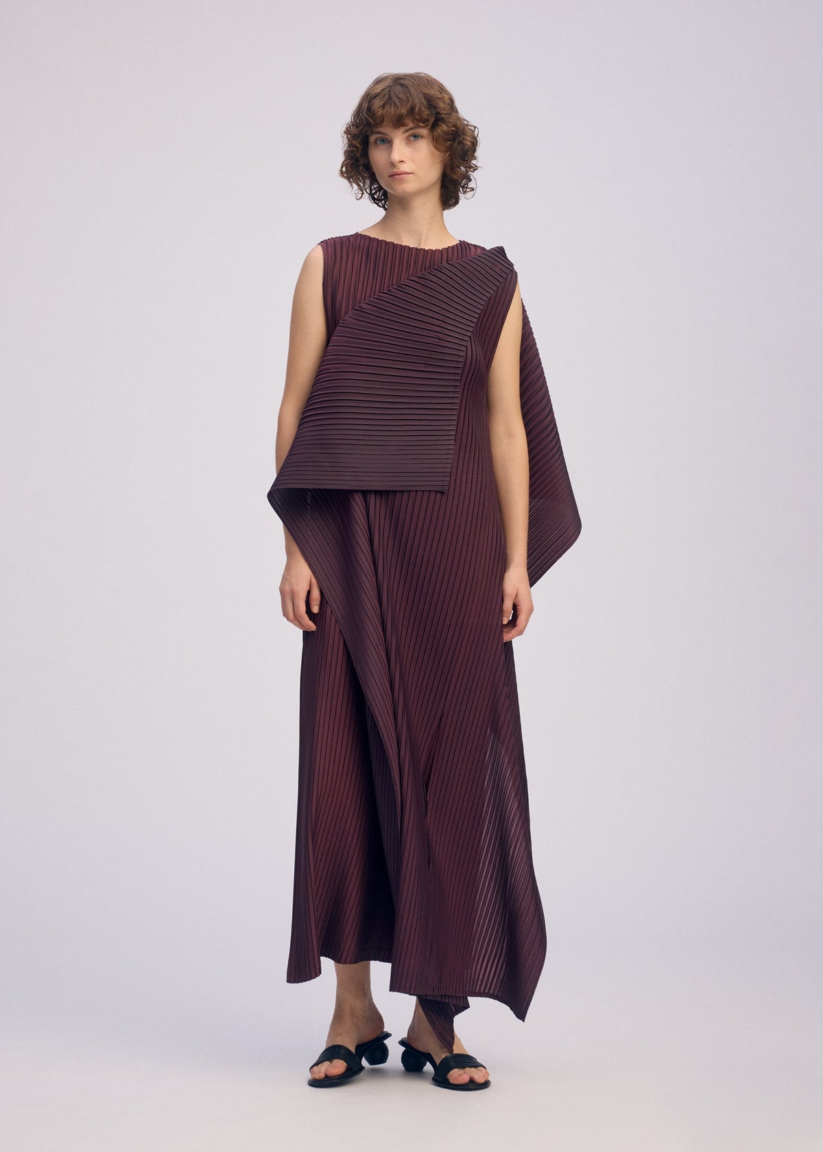 A person with short curly hair wears ISSEY MIYAKE’s PLEATED ON PLEATS—a sleeveless, asymmetric burgundy layered dress with diagonal stripes and panels—paired with black sandals against a plain light background. view 6 of 8