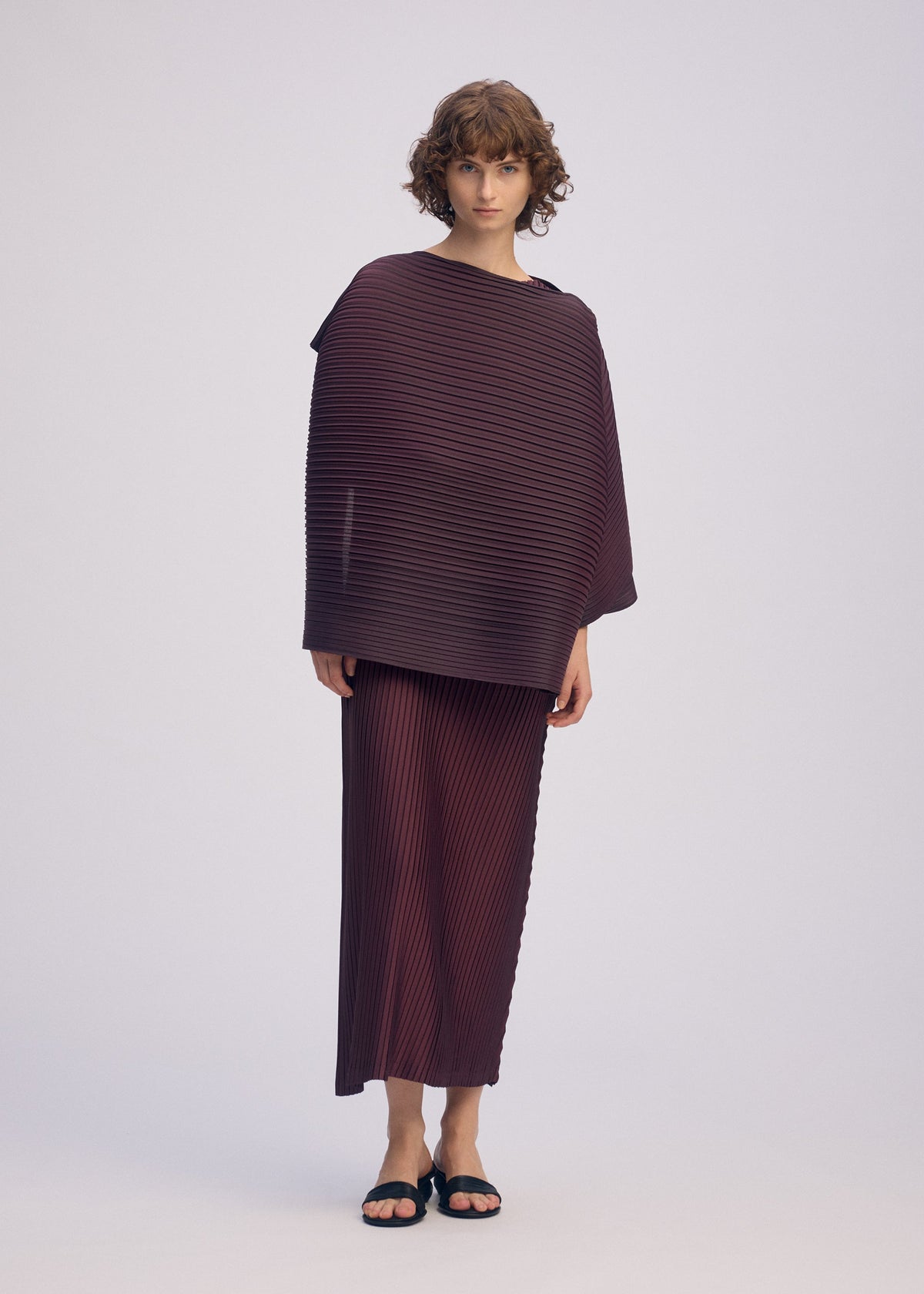 A person with short curly hair models ISSEY MIYAKE's PLEATED ON PLEATS set—an oversized burgundy ribbed top and matching skirt—styled with black sandals and a chic jacquard scarf, against a plain light background. view 7 of 8