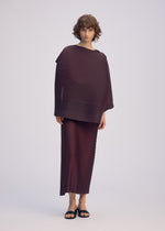 A person with short curly hair models ISSEY MIYAKE's PLEATED ON PLEATS set—an oversized burgundy ribbed top and matching skirt—styled with black sandals and a chic jacquard scarf, against a plain light background.