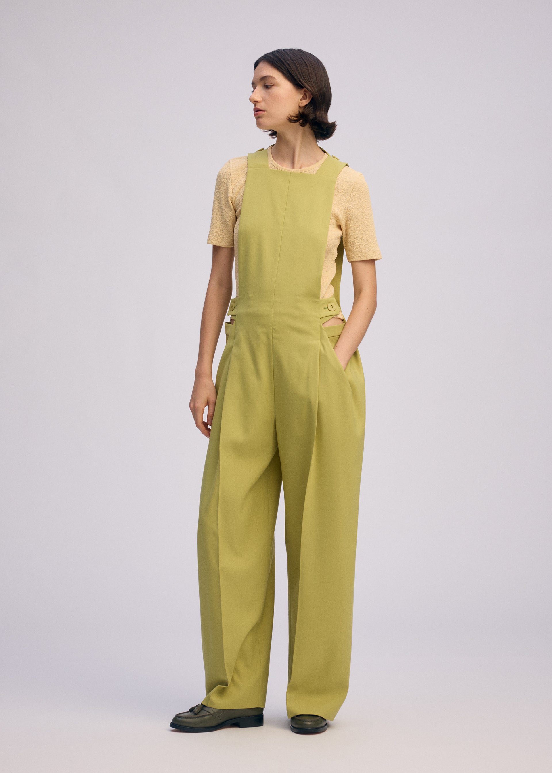 Zoomed view A person stands against a plain background wearing ISSEY MIYAKE BELTED WOOL PANTS with hands in pockets, styled with a short-sleeved beige top and dark shoes.