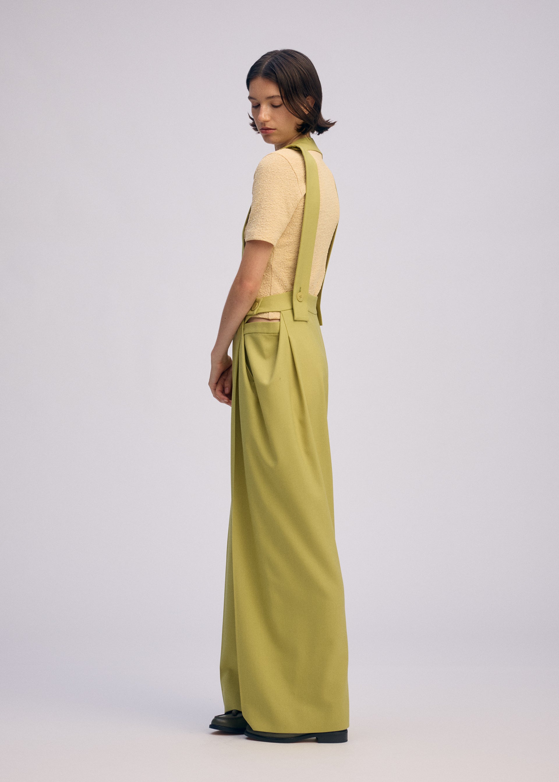 Zoomed view A person with short dark hair stands sideways, wearing a light beige top and ISSEY MIYAKE's BELTED WOOL PANTS against a plain pale background.