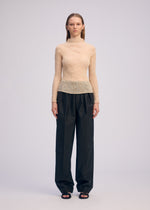 A person stands facing forward against a plain background, wearing the ISSEY MIYAKE CHIFFON TWIST 1 beige sheer long-sleeved top, loose black trousers, and black open-toe sandals. Their hair is straight and worn down.
