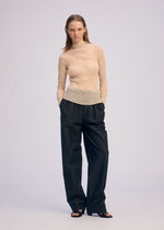 A woman stands against a plain background, wearing ISSEY MIYAKE's CHIFFON TWIST 1 sheer beige long-sleeved top, loose black trousers, and black open-toe heels. Her hands are in her pockets and she wears a neutral expression.