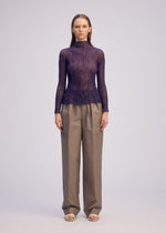 A woman stands facing forward, wearing the ISSEY MIYAKE CHIFFON TWIST 2—a sheer purple long-sleeve, high-neck top—paired with loose high-waisted beige trousers and open-toed sandals. She has straight shoulder-length hair against a plain light gray background.