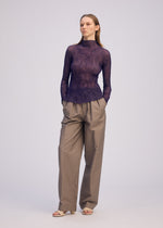 A woman stands against a plain background, wearing the ISSEY MIYAKE CHIFFON TWIST 2 sheer recycled polyester long-sleeved purple turtleneck top and loose beige pleated trousers, with her hands in her pockets and light-colored sandals.