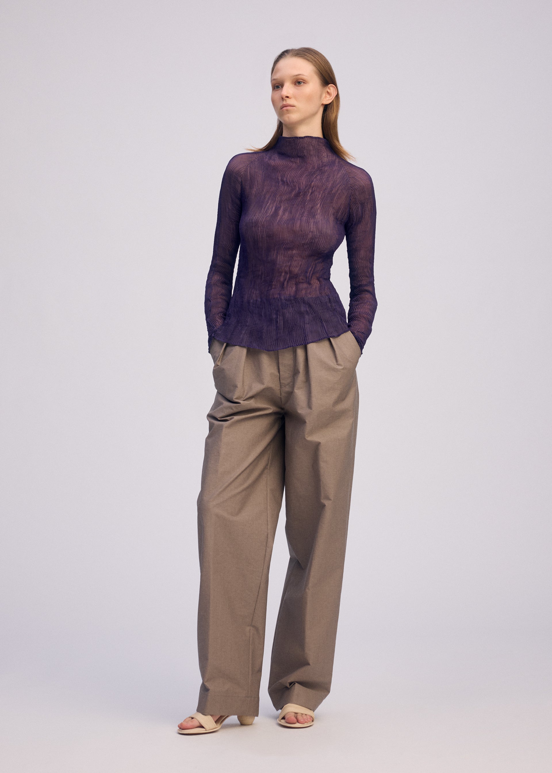 Zoomed view A woman stands against a plain background, wearing the ISSEY MIYAKE CHIFFON TWIST 2 sheer recycled polyester long-sleeved purple turtleneck top and loose beige pleated trousers, with her hands in her pockets and light-colored sandals.