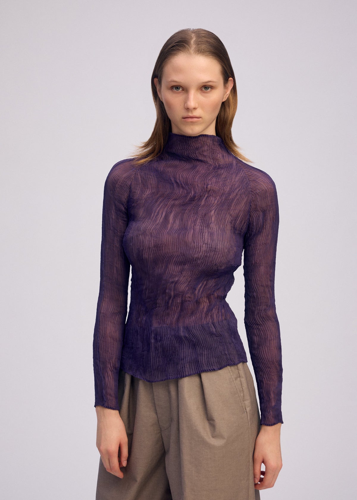 A person with straight, shoulder-length hair wears the ISSEY MIYAKE CHIFFON TWIST 2, a sheer textured purple pleated top made from recycled polyester, paired with high-waisted beige pants against a plain light background. view 7 of 8