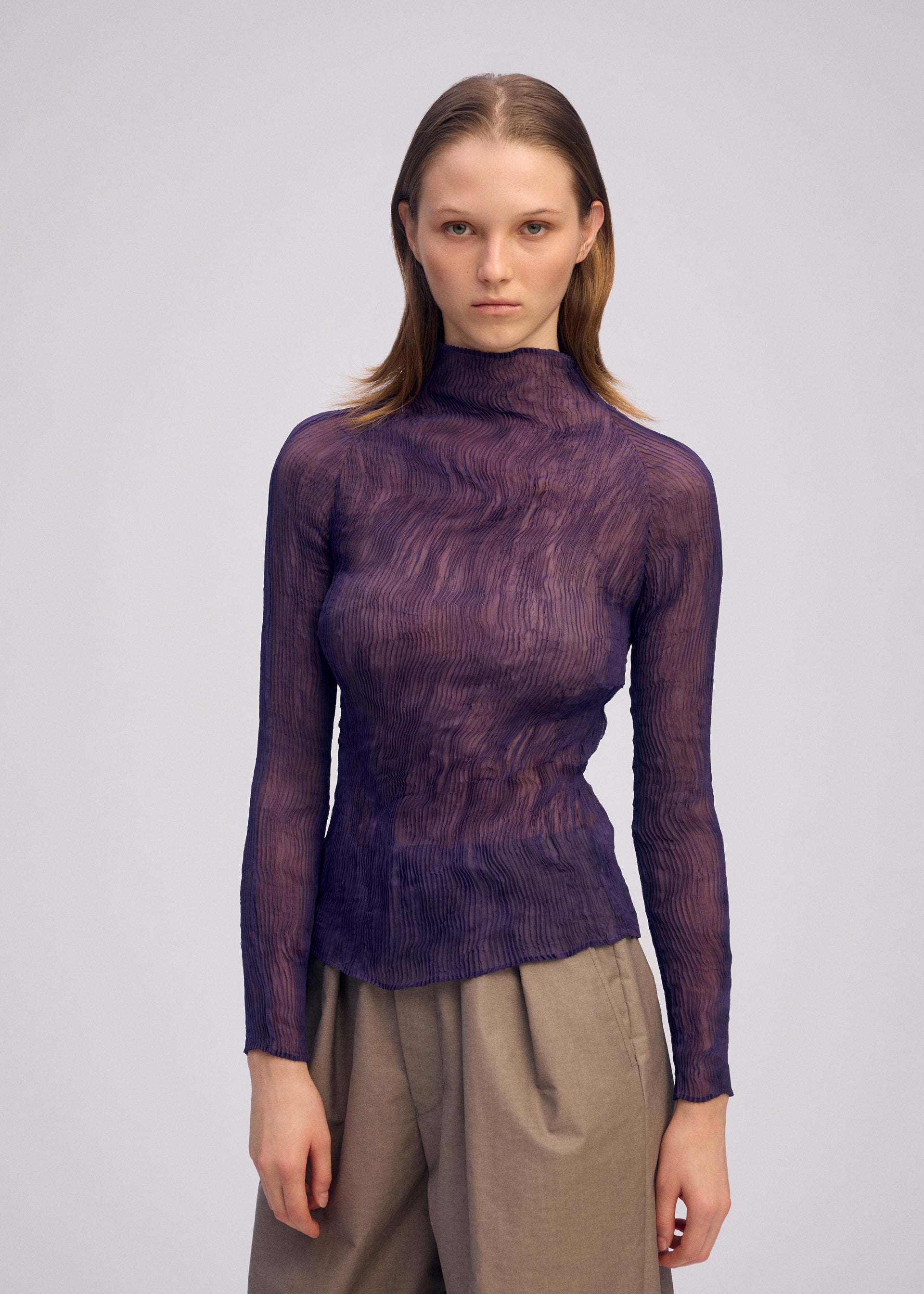 Zoomed view A person with straight, shoulder-length hair wears the ISSEY MIYAKE CHIFFON TWIST 2, a sheer textured purple pleated top made from recycled polyester, paired with high-waisted beige pants against a plain light background.