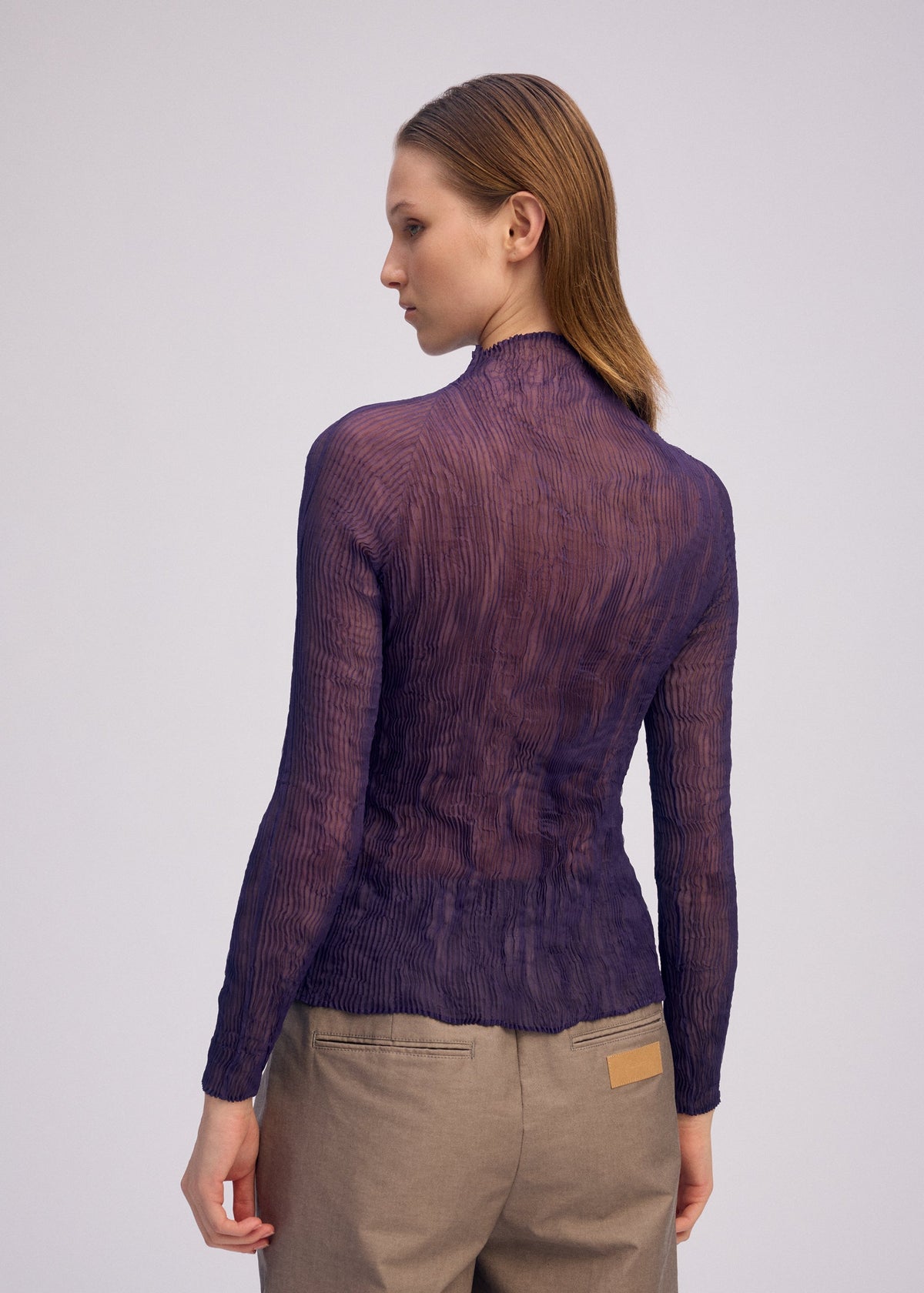 A person with straight, light brown hair is seen from behind, wearing the ISSEY MIYAKE CHIFFON TWIST 2 purple pleated sheer long-sleeve top made from recycled polyester and beige pants, standing against a plain light background. view 8 of 8
