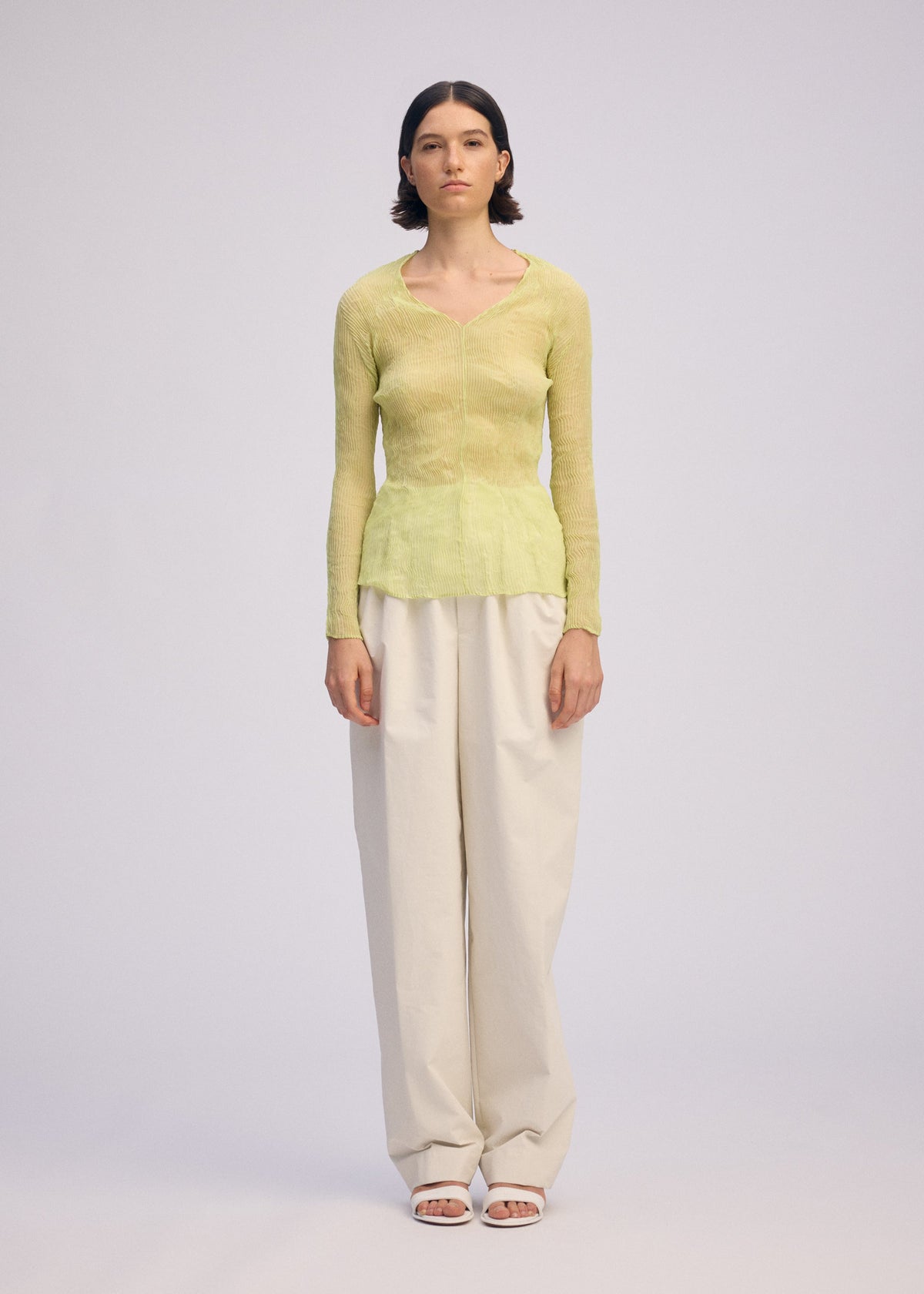 A person stands facing forward in the ISSEY MIYAKE CHIFFON TWIST 1, a sheer light green V-neck top made from recycled polyester, paired with loose off-white pants and white open-toe sandals, against a plain light gray background. view 5 of 10