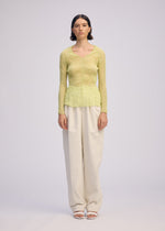 A person stands facing forward in the ISSEY MIYAKE CHIFFON TWIST 1, a sheer light green V-neck top made from recycled polyester, paired with loose off-white pants and white open-toe sandals, against a plain light gray background.