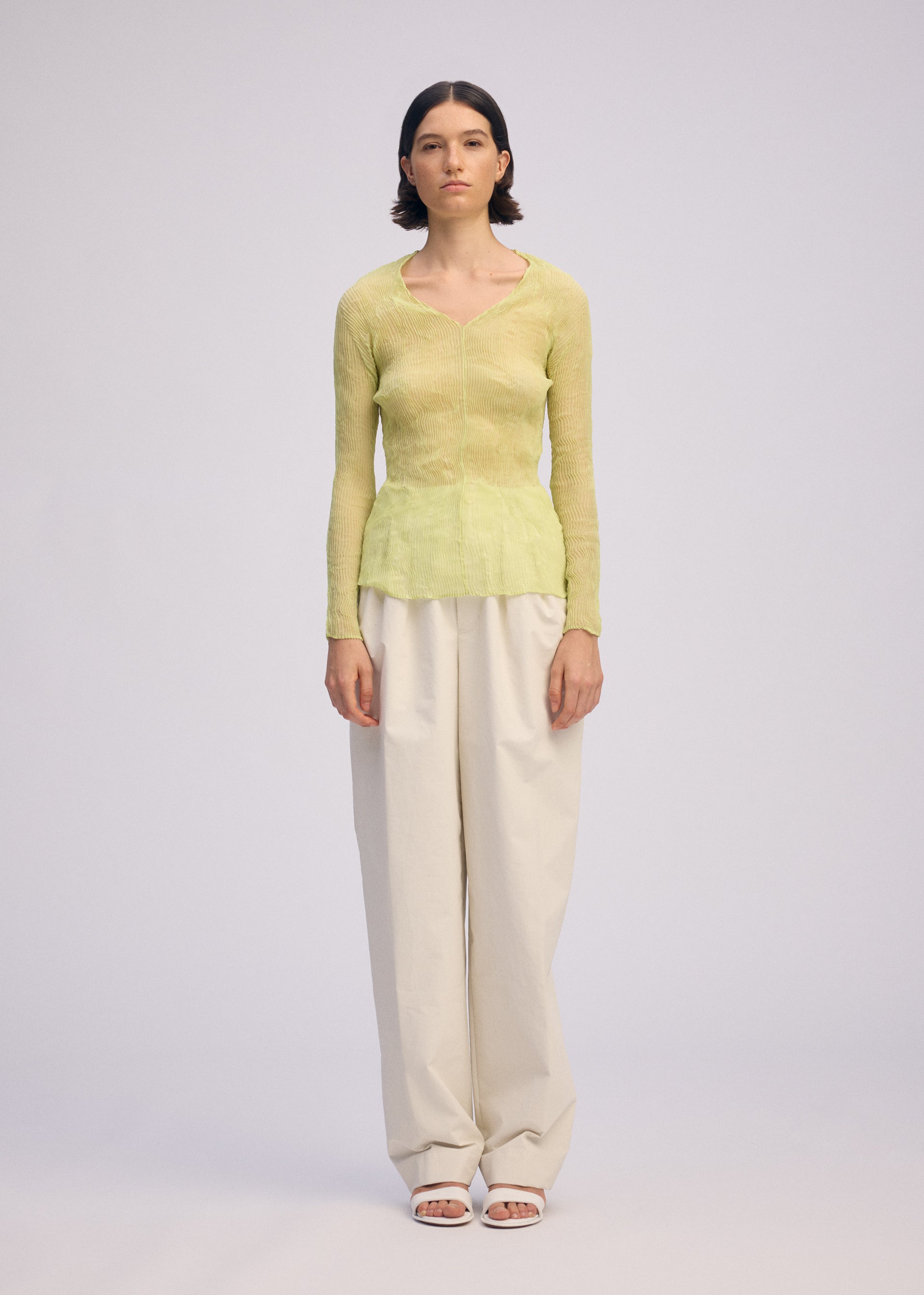 Zoomed view A person stands facing forward in the ISSEY MIYAKE CHIFFON TWIST 1, a sheer light green V-neck top made from recycled polyester, paired with loose off-white pants and white open-toe sandals, against a plain light gray background.