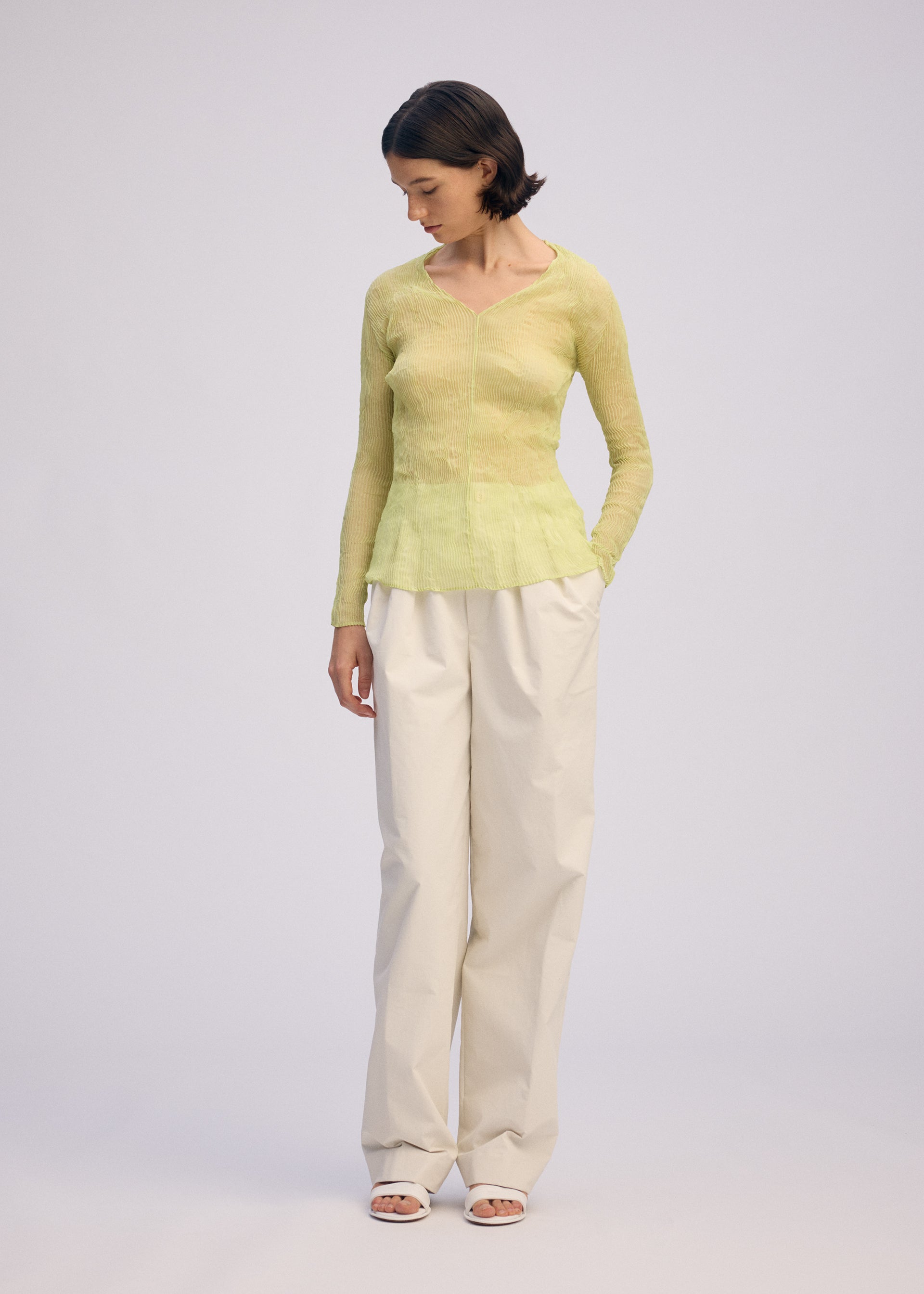 Zoomed view A person with short dark hair stands against a light background, wearing an ISSEY MIYAKE CHIFFON TWIST 1 sheer long-sleeve V-neck top in light green, off-white wide-leg pants, and white sandals, hands in pockets and looking down.