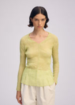 A young woman with short dark hair wears the CHIFFON TWIST 1 sheer long-sleeve textured top by ISSEY MIYAKE in light yellow, paired with white pants, standing against a plain light background.