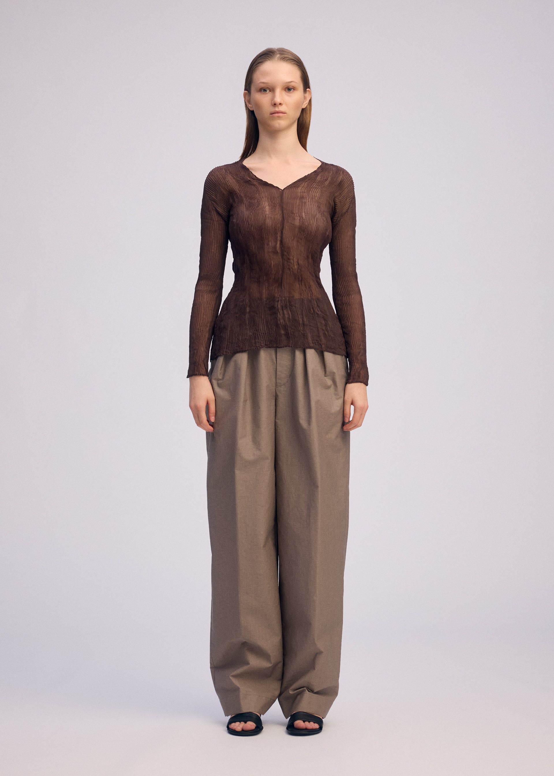 Zoomed view A person stands facing forward, wearing the ISSEY MIYAKE CHIFFON TWIST 1 sheer long-sleeve V-neck top and loose, high-waisted beige pants with black sandals against a plain light background.