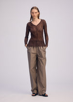 A person faces forward with hands in pockets, wearing the ISSEY MIYAKE CHIFFON TWIST 1 sheer long-sleeve V-neck top, loose beige trousers, and black sandals against a plain light background.