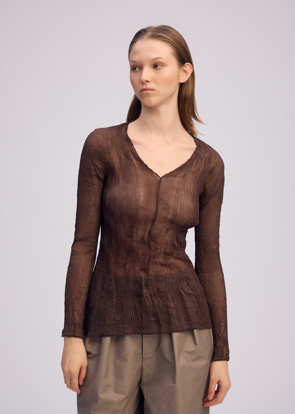 A person with straight, light brown hair wears the ISSEY MIYAKE CHIFFON TWIST 1 sheer long-sleeve V-neck top and loose taupe pants, posing against a plain light background and gazing slightly to the side. view 10 of 10