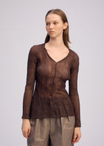 A person with straight, light brown hair wears the ISSEY MIYAKE CHIFFON TWIST 1 sheer long-sleeve V-neck top and loose taupe pants, posing against a plain light background and gazing slightly to the side.