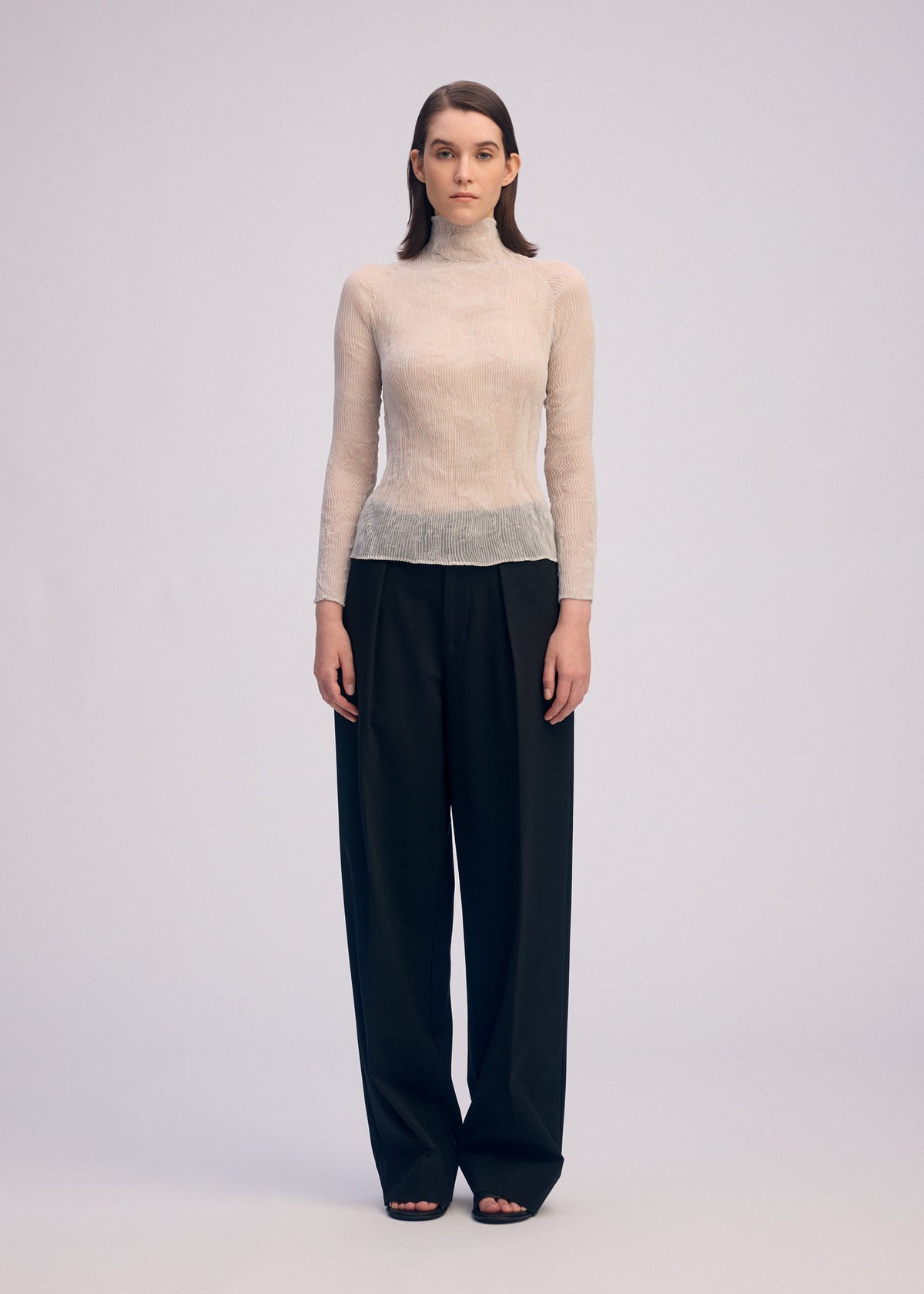 A person stands facing forward, wearing the ISSEY MIYAKE CHIFFON TWIST 3 sheer long-sleeved top with loose, high-waisted black pants against a plain light background for a minimalist look. view 6 of 8