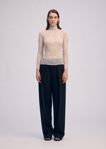 A person stands facing forward, wearing the ISSEY MIYAKE CHIFFON TWIST 3 sheer long-sleeved top with loose, high-waisted black pants against a plain light background for a minimalist look.