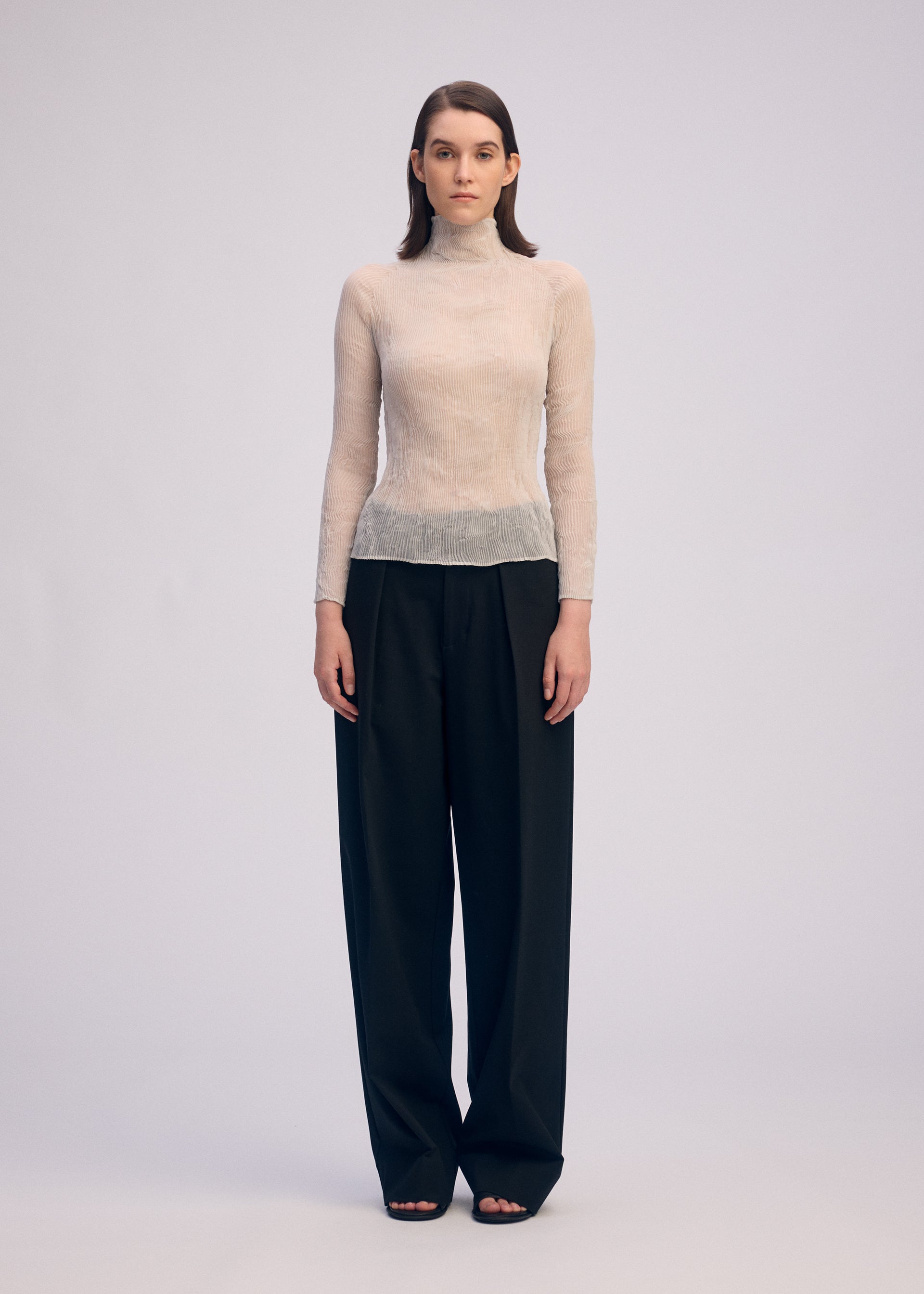 Zoomed view A person stands facing forward, wearing the ISSEY MIYAKE CHIFFON TWIST 3 sheer long-sleeved top with loose, high-waisted black pants against a plain light background for a minimalist look.