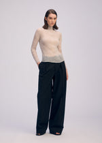 A woman with straight, shoulder-length brown hair and a neutral expression models ISSEY MIYAKE’s CHIFFON TWIST 3: a sheer, long-sleeved top made from recycled polyester, paired with high-waisted wide-leg black trousers and open-toe black heels.