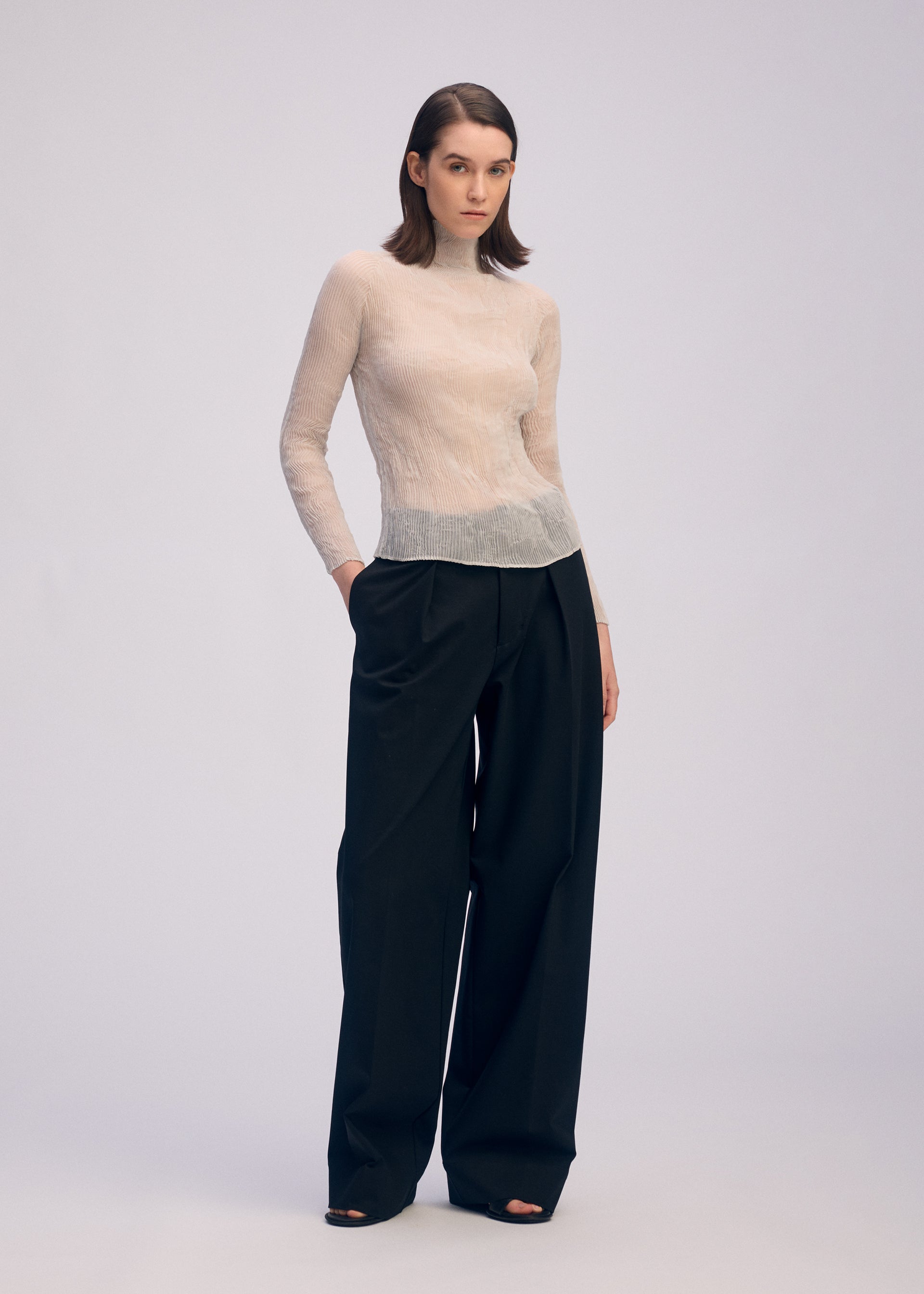 Zoomed view A woman with straight, shoulder-length brown hair and a neutral expression models ISSEY MIYAKE’s CHIFFON TWIST 3: a sheer, long-sleeved top made from recycled polyester, paired with high-waisted wide-leg black trousers and open-toe black heels.