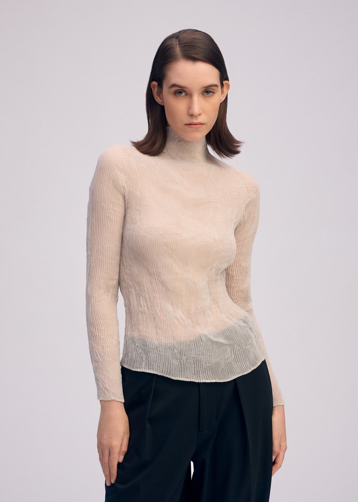 A woman with straight brown hair wears ISSEY MIYAKE's CHIFFON TWIST 3 sheer recycled polyester top and loose black trousers, standing against a light background and looking directly at the camera with a neutral expression. view 8 of 8