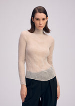 A woman with straight brown hair wears ISSEY MIYAKE's CHIFFON TWIST 3 sheer recycled polyester top and loose black trousers, standing against a light background and looking directly at the camera with a neutral expression.