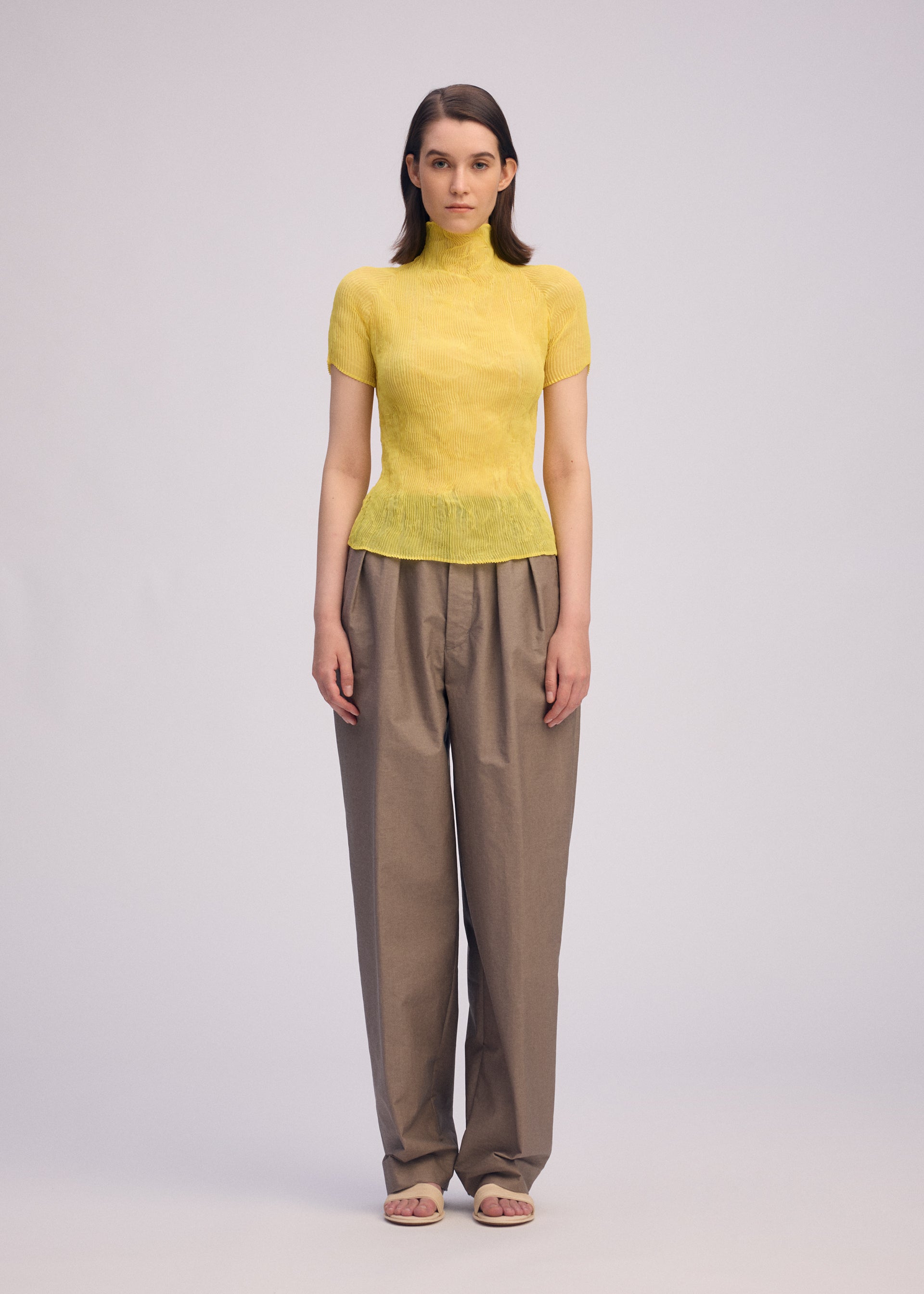 Zoomed view A person stands facing forward against a plain light background, wearing the ISSEY MIYAKE CHIFFON TWIST 3 fitted short-sleeve yellow pleated top, loose tan trousers, and beige open-toe sandals.
