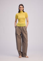 A woman stands with hands in pockets, wearing the ISSEY MIYAKE CHIFFON TWIST 3 yellow pleated top, loose brown wide-leg trousers, and beige sandals against a plain light background.