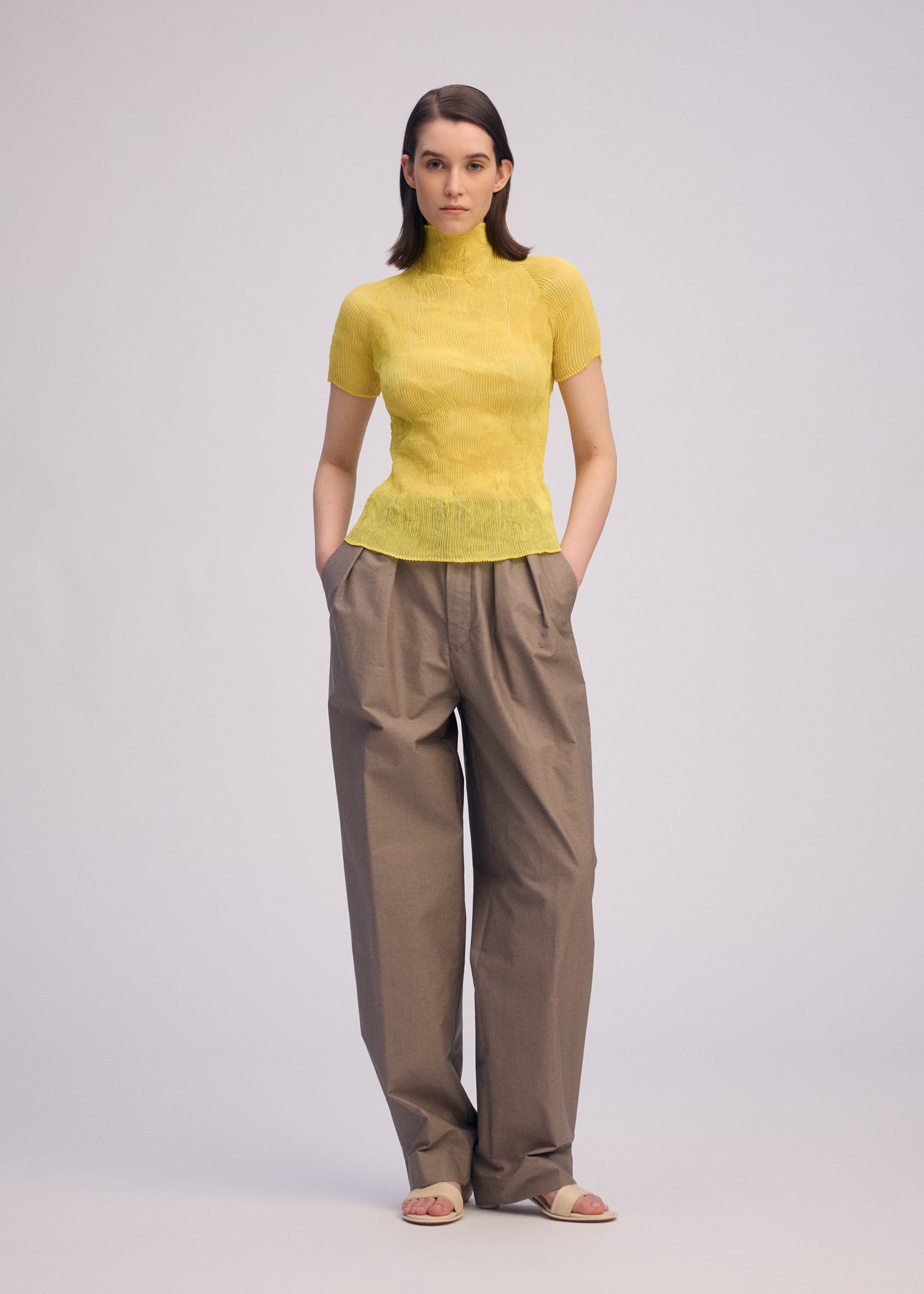 Zoomed view A woman stands with hands in pockets, wearing the ISSEY MIYAKE CHIFFON TWIST 3 yellow pleated top, loose brown wide-leg trousers, and beige sandals against a plain light background.