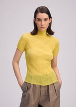 A person with straight, shoulder-length brown hair wears the ISSEY MIYAKE CHIFFON TWIST 3 pleated yellow top and loose brown pants, standing with hands in pockets against a plain light background.