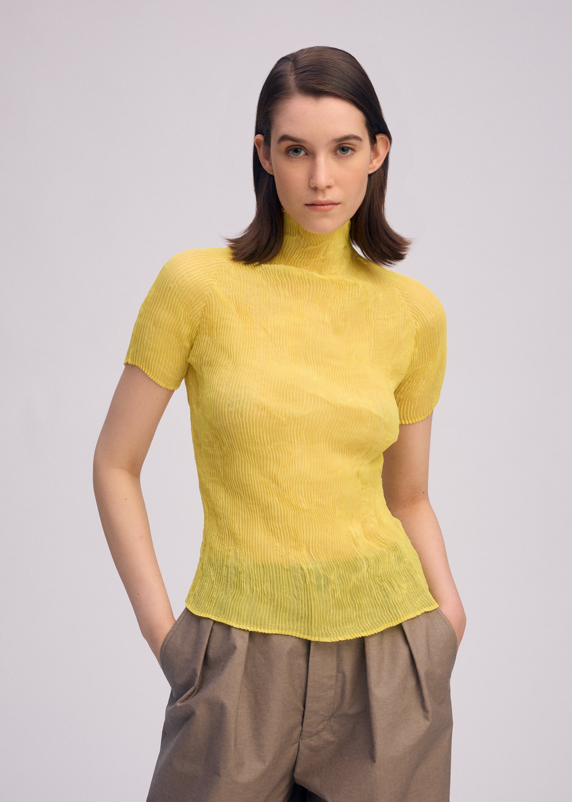 Zoomed view A person with straight, shoulder-length brown hair wears the ISSEY MIYAKE CHIFFON TWIST 3 pleated yellow top and loose brown pants, standing with hands in pockets against a plain light background.