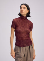 A woman with short dark hair models the ISSEY MIYAKE CHIFFON TWIST 3, a textured maroon sheer high-neck top paired with loose beige pants, standing against a plain light background and gazing slightly to the side.