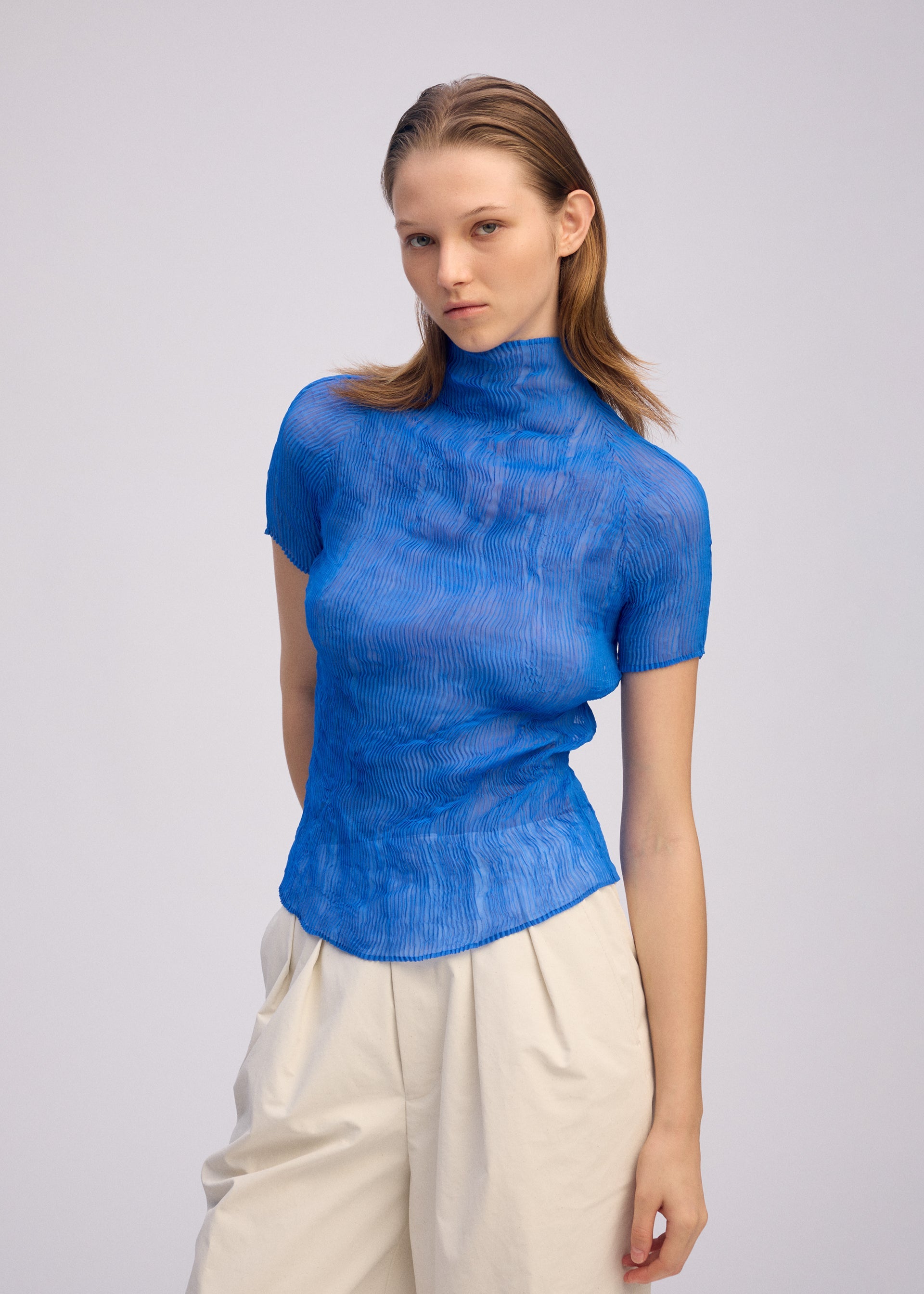 Zoomed view A woman with straight, light brown hair wears the ISSEY MIYAKE CHIFFON TWIST 3 pleated, semi-sheer blue top with high-waisted off-white pants. She stands against a plain light background, looking at the camera with a neutral expression.