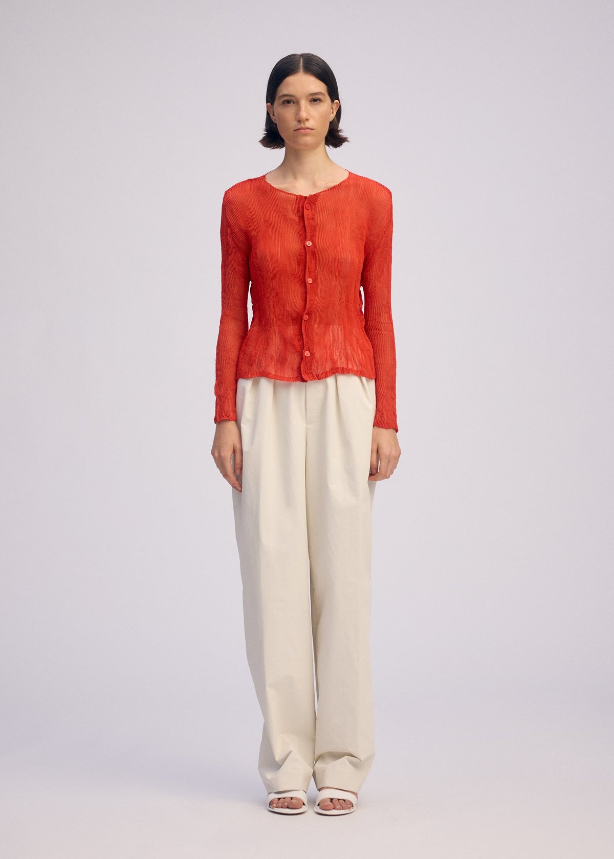 A woman with short dark hair stands against a plain background, wearing ISSEY MIYAKE's CHIFFON TWIST 2—a long-sleeved red button-up blouse—with high-waisted loose white recycled polyester trousers and white open-toe sandals. view 5 of 9