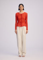 A woman with short dark hair stands against a plain background, wearing ISSEY MIYAKE's CHIFFON TWIST 2—a long-sleeved red button-up blouse—with high-waisted loose white recycled polyester trousers and white open-toe sandals.