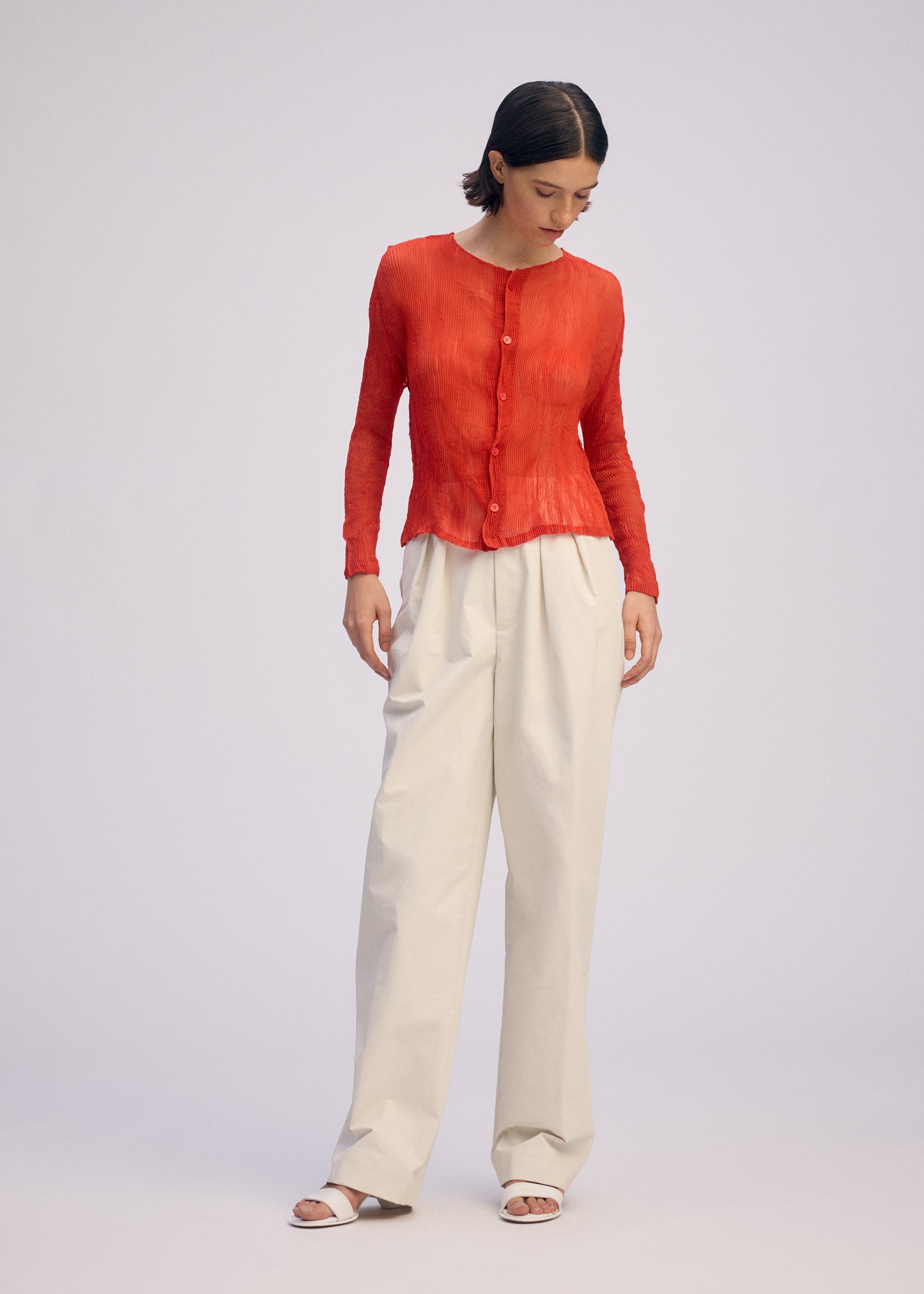 Zoomed view A woman stands against a plain background wearing ISSEY MIYAKE's CHIFFON TWIST 2, paired with loose off-white trousers and white sandals. She looks down, hands relaxed by her sides.