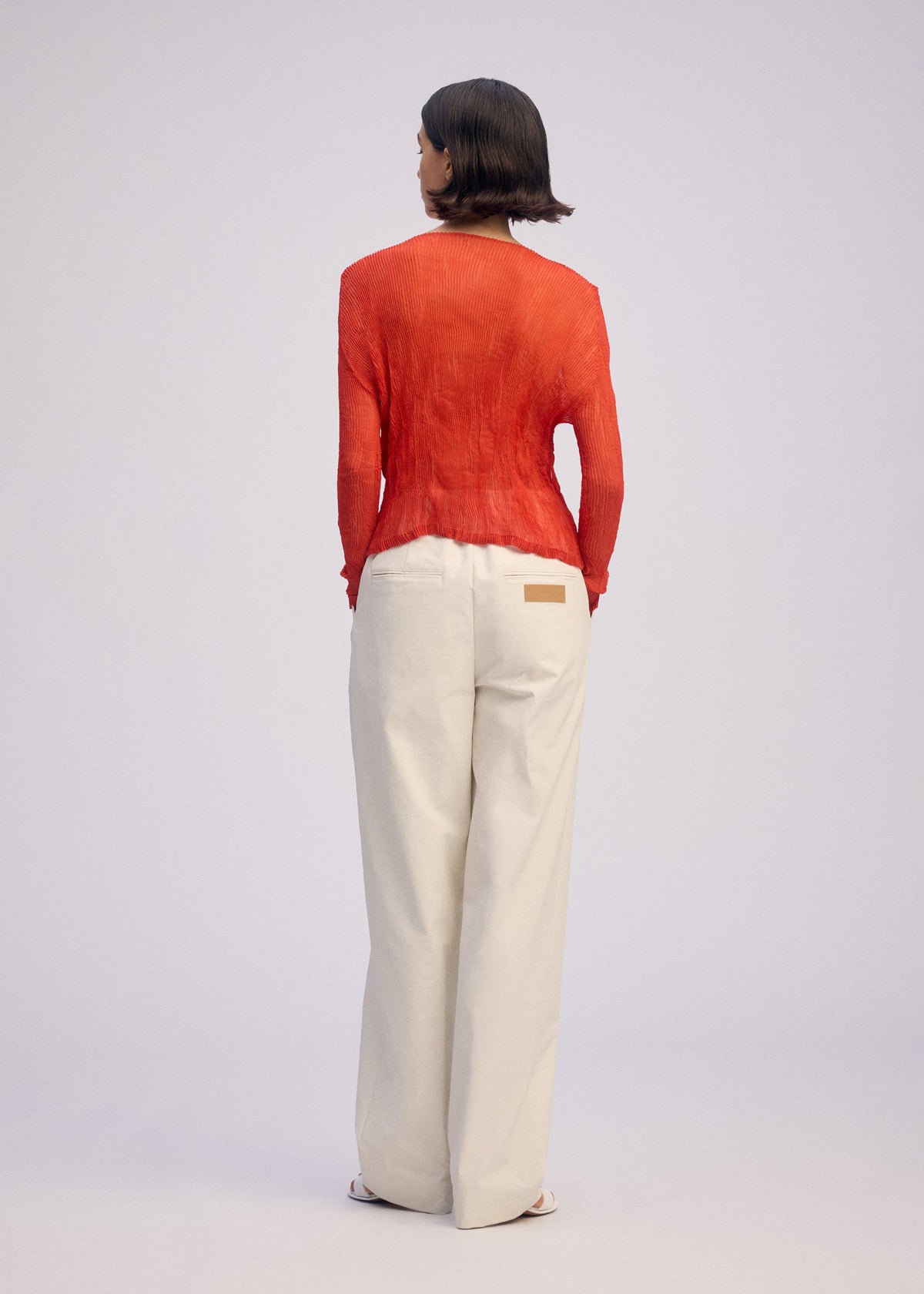 A person with short brown hair faces away, wearing the ISSEY MIYAKE CHIFFON TWIST 2 long-sleeved red sheer cardigan and loose white pants with a tan waistband patch against a plain light background. view 7 of 9