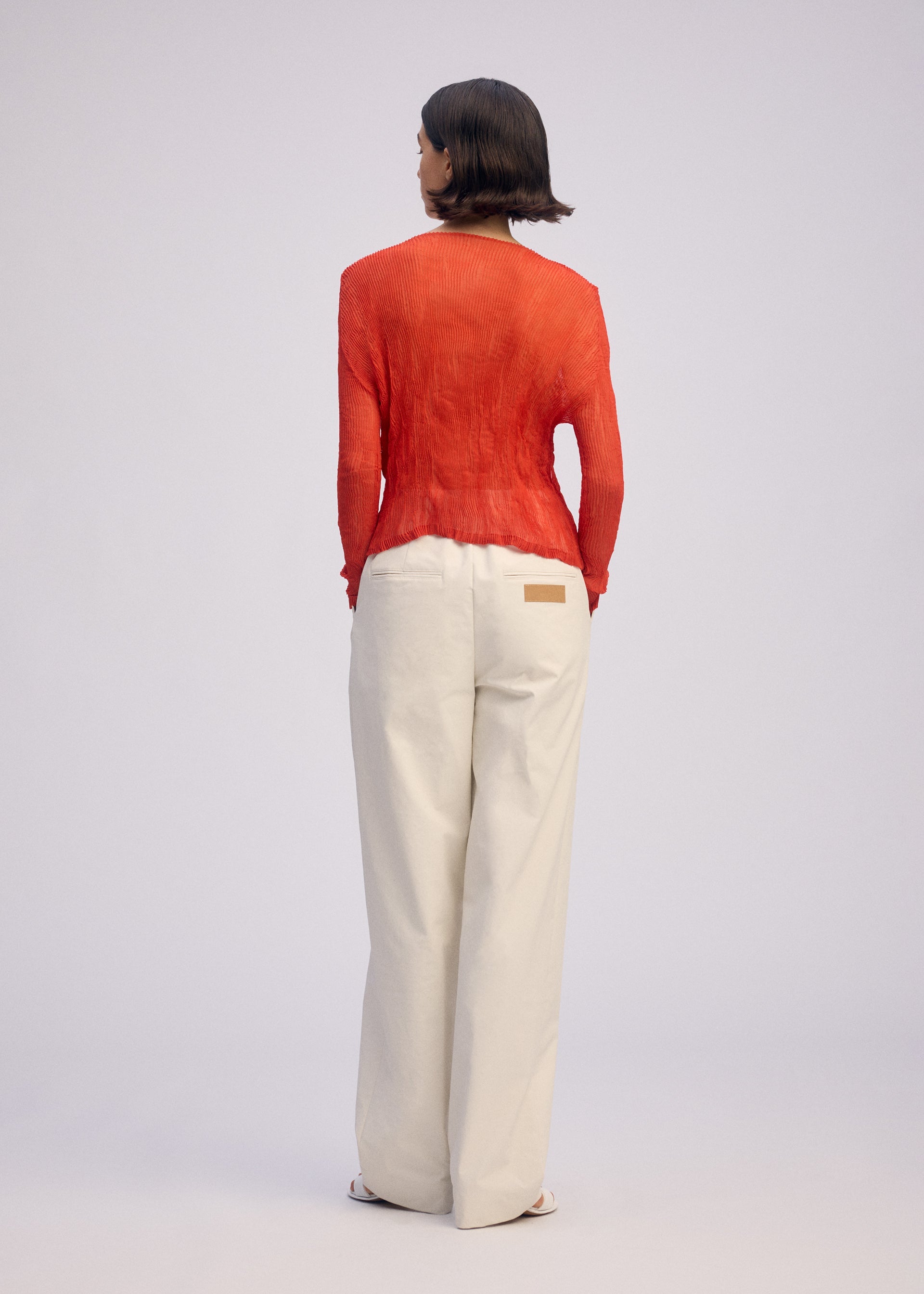 Zoomed view A person with short brown hair faces away, wearing the ISSEY MIYAKE CHIFFON TWIST 2 long-sleeved red sheer cardigan and loose white pants with a tan waistband patch against a plain light background.