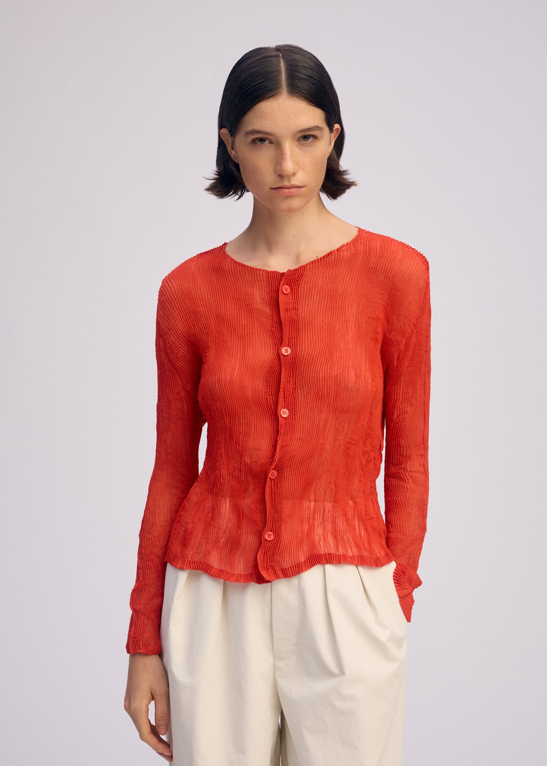 Zoomed view A person with short dark hair wears the ISSEY MIYAKE CHIFFON TWIST 2, a textured red long-sleeved pleated cardigan, and high-waisted loose white pants, standing against a plain light background.